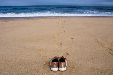 Pink canvas shoes resting on the sand facing the ocean at empty beach with footprints and waves in the background. Girl took off shoes and left footprints in the sand.