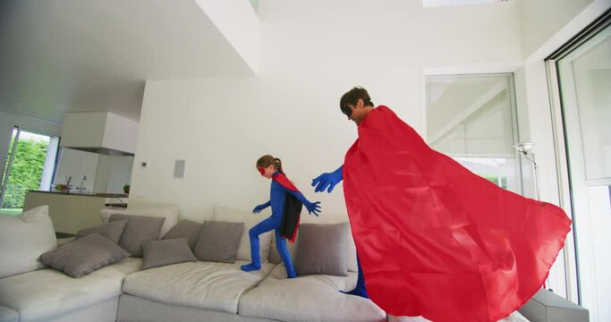 A carefree happy smiling father and daughter dressed as superheroes are having fun to run and jump while playing an imagination game at home.
