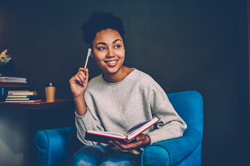 Smiling creative female blogger pondering on idea for publication sitting in comfortable armchair,young african american woman recalling information while planning to do list at home interior  . - obrazy, fototapety, plakaty