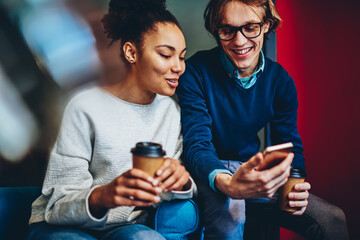 Cheerful diversity friends watching funny video on smartphone resting together on coffee break, positive male and female colleagues talking to each other reading information fron cellphone in office