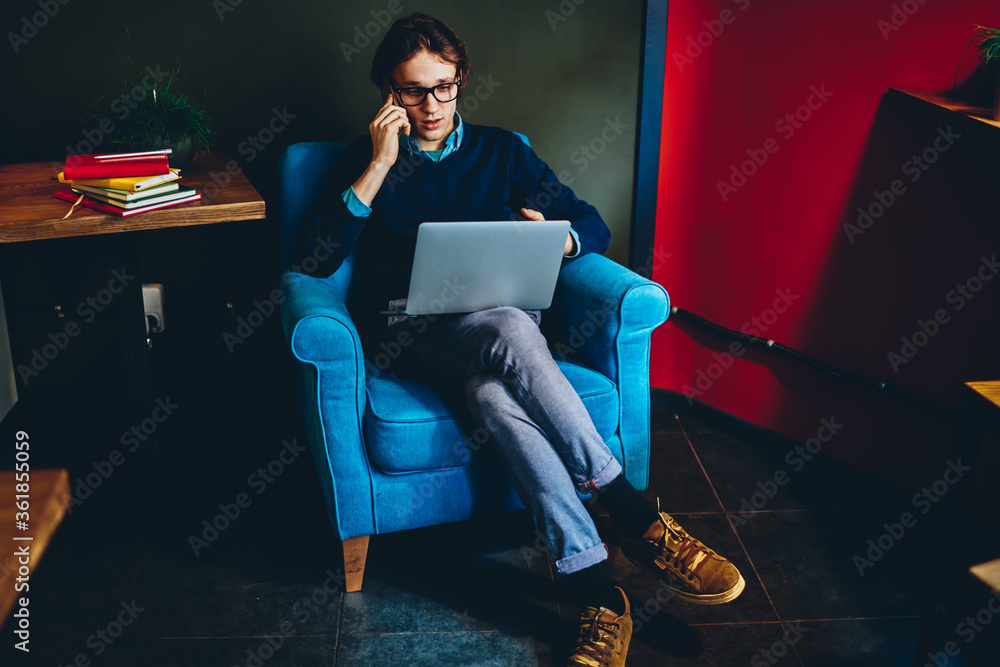 Aufkleber young hipster guy talking on mobile phone while using netbook for remote job at home, serious male c