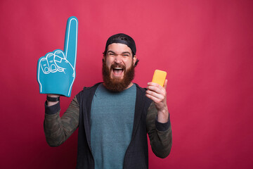 Excited bearded male hipster is wearing blue fan foam glove and his phone screaming at the camera