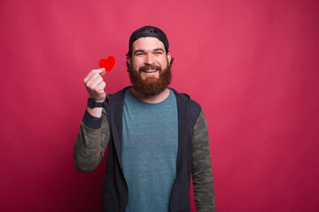 Smiling at the camera hipster man is holding a red little heart over pink background