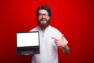 Happy bearded man wearing glasses is pointing at a blank screen of a laptop over red background.