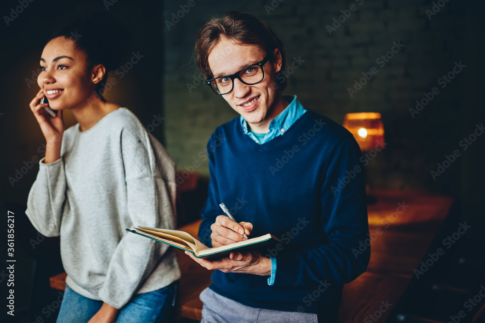 Poster skilled male in eyewear making notes in notepad looking at camera during working process in office w