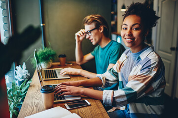 Cheerful dark skinned female freelancer satisfied with online business working with male colleague on netbooks, positive multiracial friends learning via laptop computers  and apps in cafe interior