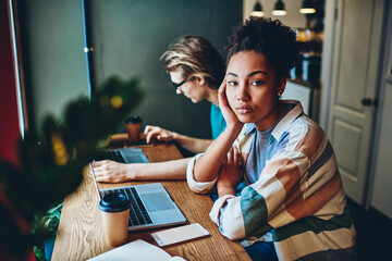 Serious dark skinned woman looking at camera while her colleague concentrated on remote job via laptop computer, multiracial friends using wireless connection in coworking space for online work.