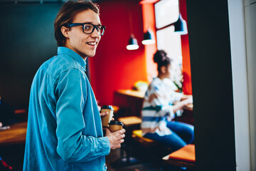 Positive caucasian man in eyewear carrying coffee to female friend spending time in cafe interior in morning, rear view of cheerful hipster guy holding cardboard cups with beverage in cafe interior