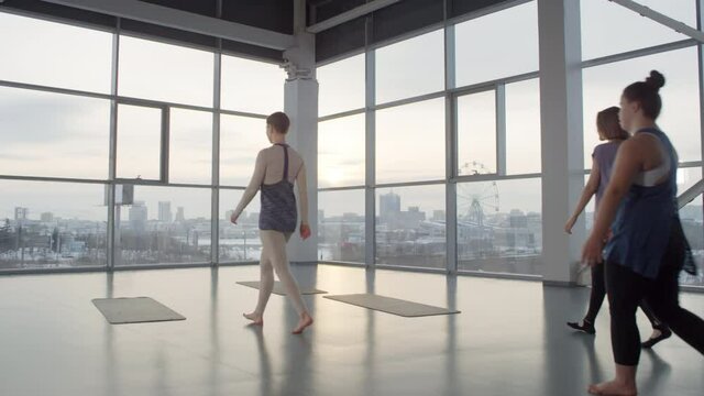 Tracking backlit long shot of trainer and two women coming in spacious yoga hall with panoramic windows in early morning to workout