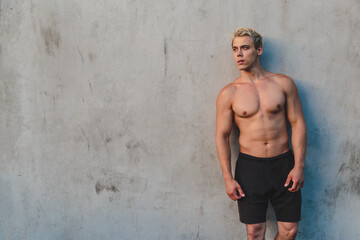 Natural portrait of young athletic shirtless man standing near concrete wall.