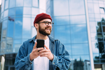 Portrait of happy handsome bearded young male hipster holding smartphone and looking to the side - Powered by Adobe