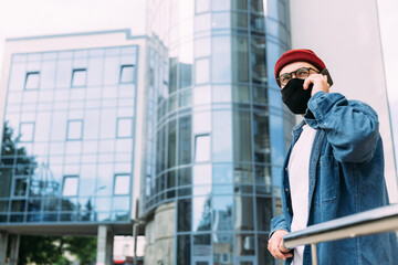 Bearded male hipster in protective mask talking on mobile phone in business district