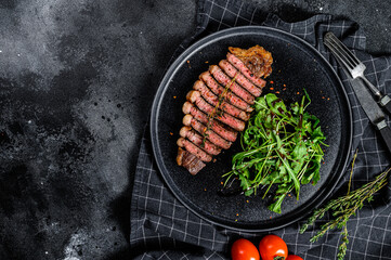 Sliced sirloin steak, marbled beef meat with arugula. Black background. Top view. Copy space