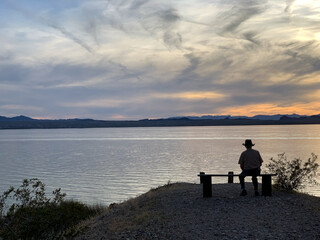 Silhouette of a man sitting on a bench overlooking a lake at sunset 