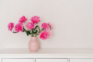 big bouquet of fuchsia peonies in a pink vase in a bright room are on a white chest of drawers. cozy house, fresh flowers at home, home greenhouse, minimal design, loft house