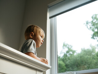 Boy toddler looks out the window from crib on a cloudy day