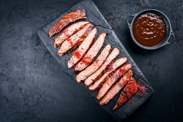 Traditional barbecue sliced dry aged wagyu flank steak offered with spicy BBQ Sauce and chili powder as overhead view on a rustic charred wooden cutting board with copy space left