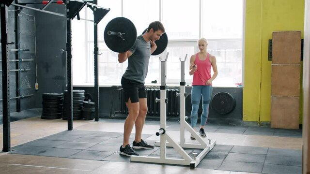 Muscular young man training in gym squatting with barbell working out with coach attractive girl wearing sports clothing. People, workout and leisure concept.