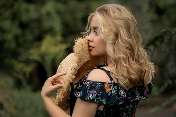 Naklejka premium A beautiful blond girl holds a straw hat near her face and looks away. A woman stands with her back to the frame. She has beautiful blue eyes