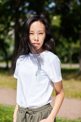 Portrait of attractive and confident Asian young woman in white t-shirt looking at camera standing outdoors in park on warm summer day