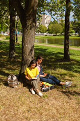 Two female college students sitting in shade of tree leaning on trunk in park. Asian girl reading textbook, her Caucasian friend using laptop. Backpacks, binders and vr headsets near them