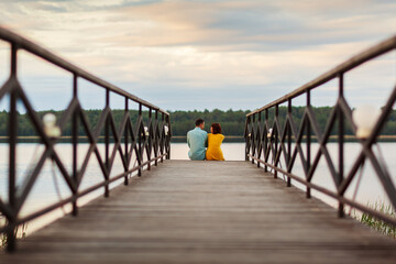 Rear view of romantic young couple sitting on wooden pier over river, enjoying warm summer evening in nature and talking