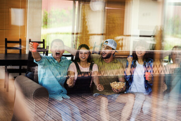 Through window glass shot of four young friends sitting on sofa at home, watching sports game on tv and screaming happily celebrating goal