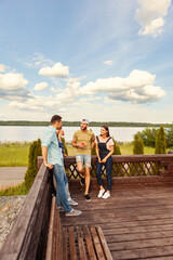 Group of four young friends, two men and two women, standing on wooden terrace in countryside with river and cloudy sky in background and enjoying talking