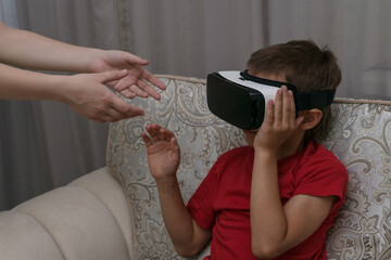 female hands want to take virtual reality glasses  from boy's eyes. Boy is sitting in big armchair and watch something interesting in cyberspace. Impact of modern technologies for children.