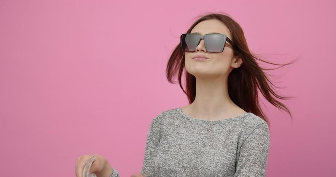 Arrogant young woman in trendy sunglasses holding fan made of dollars banknotes while posing in studio. Beautiful girl with dark hair enjoying rich life over pink background.