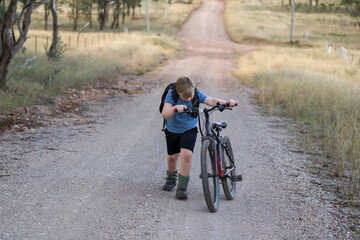 Young boy pushing bike up hill on gravel road in dry countryside