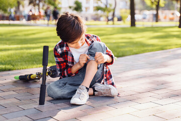 A little boy 8 years riding scooter and fell. Sitting on asphalt, holding hands on his injured leg and crying in pain. Broking his knee. It's an unexpected incident. Summer walks in the park.
