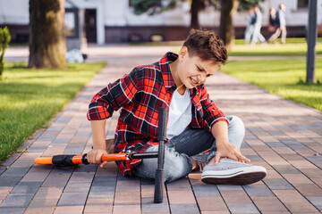 A little boy 8 years riding scooter and fell. Sitting on asphalt, holding hands on his injured leg and crying in pain. Broking his knee. It's an unexpected incident. Summer walks in the park.