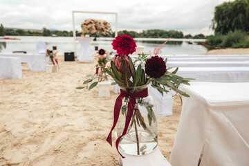 Dark red dahlia flowers in transparent glass vase. Outdoor wedding ceremony on sandy beach. Wedding day concept.