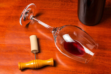 An overturned glass of wine with a little red wine next to a corkscrew and a bottle on a wooden bottom.