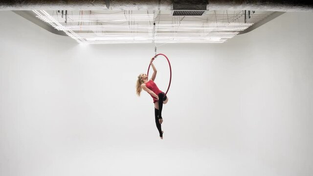 Flexible pretty blonde girl in a red dress and black
stockings performs acrobatic elements in an air hoop,
white cyclorama location.