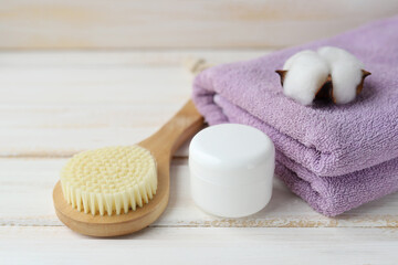 Fototapeta na wymiar Spa treatments in the sauna. A wooden massage brush for cellulite prevention, a white jar with skin care product, a purple Terry towel and a cotton flower on a white wooden background. Selective focus