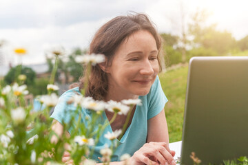 young smiling woman lying on grass with laptop in park on sunny day, remote working