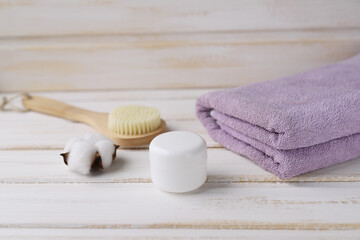 Fototapeta na wymiar Spa procedures. Wooden massage brush for cellulite prevention, white jar with skin care product, purple Terry towel and cotton flower on a white wooden background. Selective focus