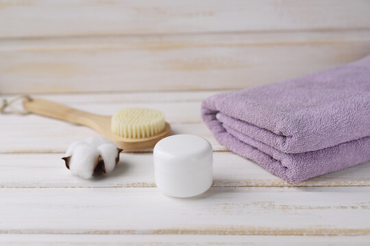 Spa procedures. Wooden massage brush for cellulite prevention, white jar with skin care product, purple Terry towel and cotton flower on a white wooden background. Selective focus