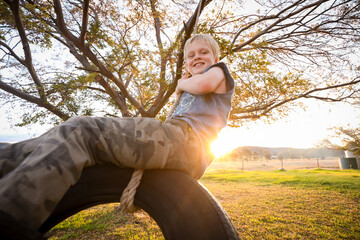 Happy little blonde boy playing and swinging on tire swing under chinese elm tree during vibrant sunset