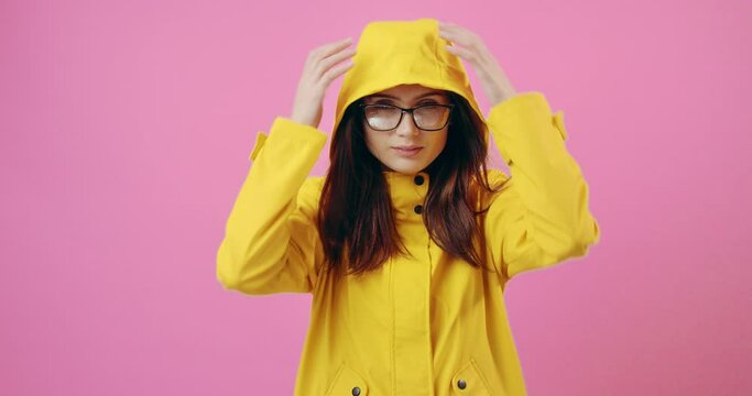 Beautiful young woman in eyeglasses and yellow raincoat with hood posing on camera with pink background. Concept of rain and bad weather.