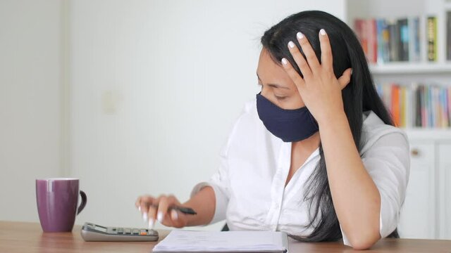 A woman sitting at a desk and wearing a face mask is looking at her finances and getting very worried and frustrated
