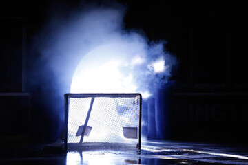 An ice hockey goal standing in the shadow in front of the player entrance with bright light and smoke coming out.