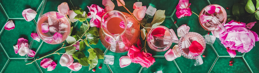 Summer refreshing cold beverage drink. Flat-lay of rose lemonade with ice in glasses and rose flower petals over green tile table background, top view, wide composition