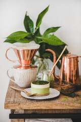 Coffee and dessert setup. Green matcha cheesecake and brewed black coffee in copper pot on rustic wooden kitchen counter, white wall background, close-up. Healthy, vegan, vegetarian, low calorie food
