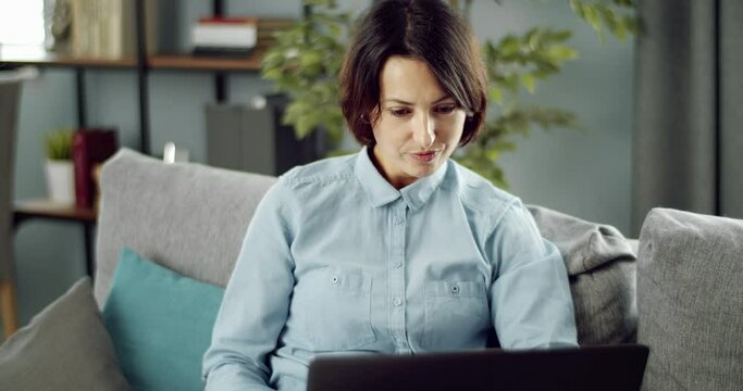 Attractive lady with short haircut sitting comfortably on sofa and browsing internet on wireless laptop. Mature lady in casual clothing using computer during free time at home.