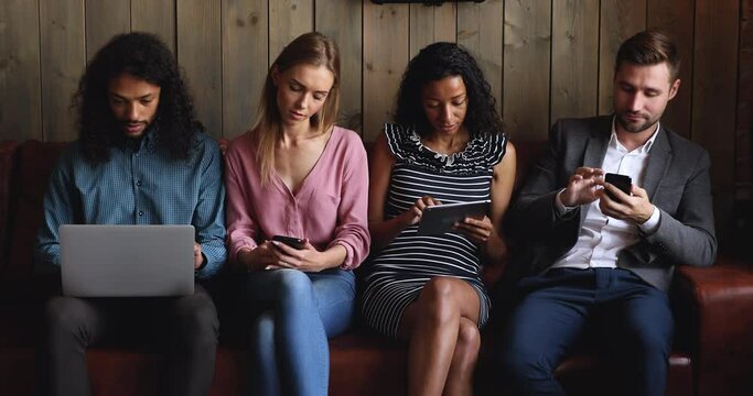 Technology addicted young multiracial people group lost in gadgets holding laptop digital tablet phones overuse social media networks sitting in row, diverse users ignoring each other obsessed with