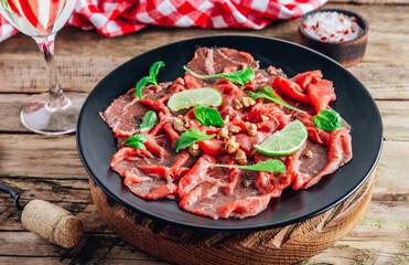Black plate with carpaccio of beef with baby arugula, lime and white wine on rustic wooden background.