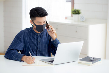 Young asian business man in face mask talking smartphone and work on laptop computer for protect covid-19, businessman quarantine work from home and writing notebook, social distancing and stay home.